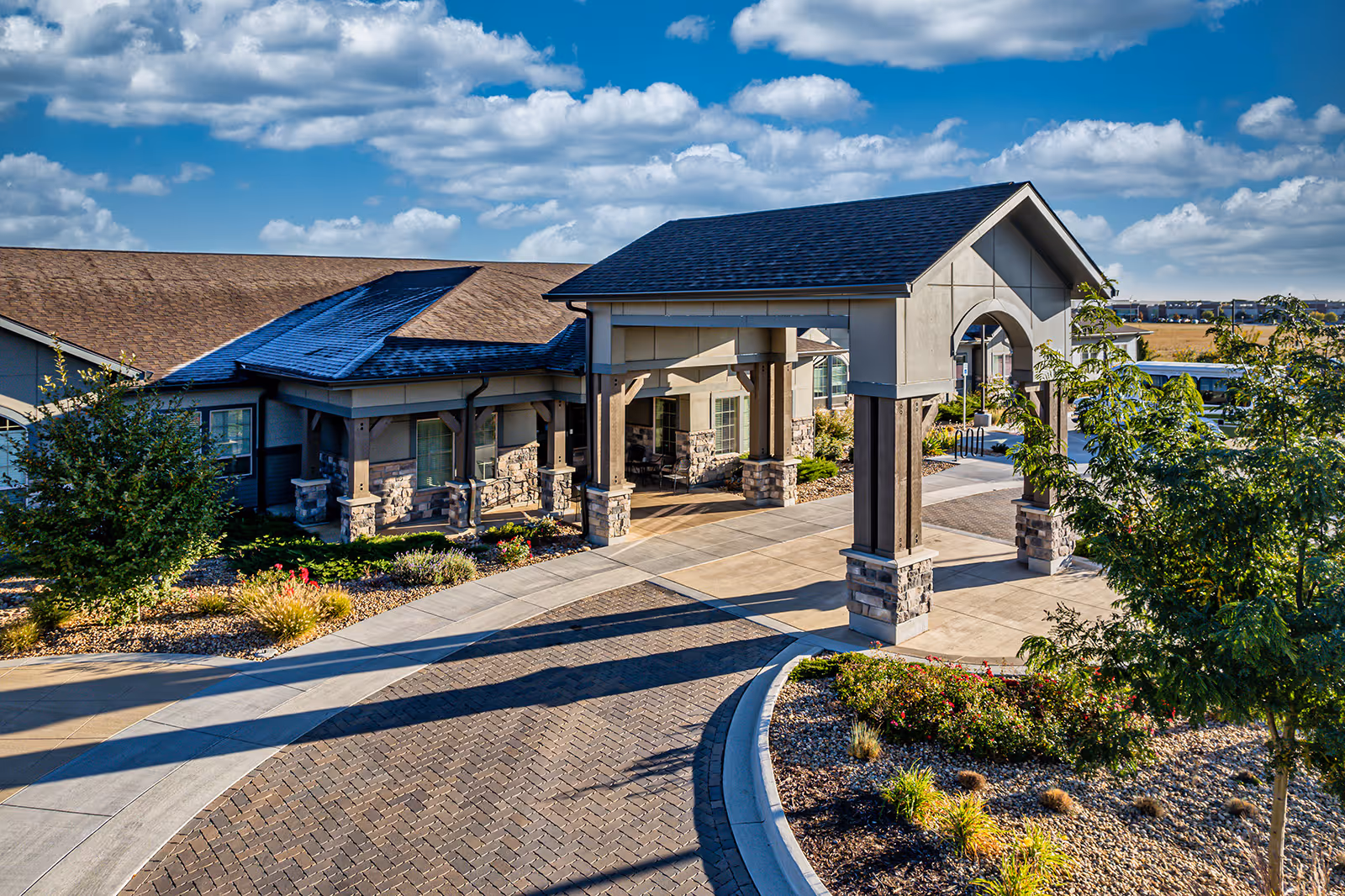 Exterior view of a senior living facility with a covered entrance supported by stone and wooden pillars, landscaped garden beds with shrubs and flowers, a paved driveway, and a blue sky with scattered clouds.