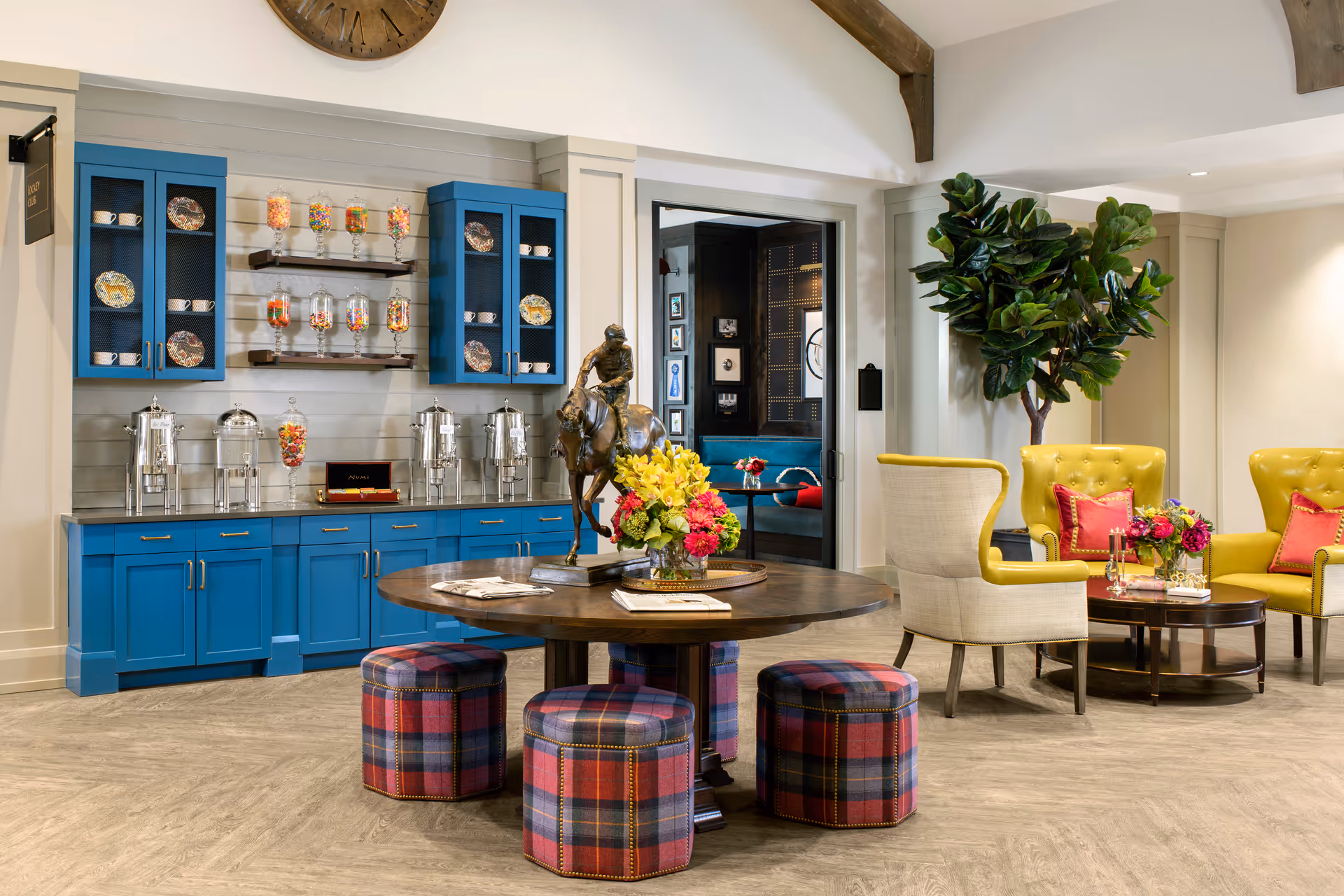 A bright and inviting common area with blue cabinets and shelves holding glass jars filled with colorful candy. A round wooden table with a bronze horse statue and flower arrangements is surrounded by four plaid upholstered stools. To the right, there are yellow armchairs with red pillows and a small round coffee table with flowers. A large potted plant is in the corner, and a doorway leads to another room with a blue sofa and framed pictures on the wall.