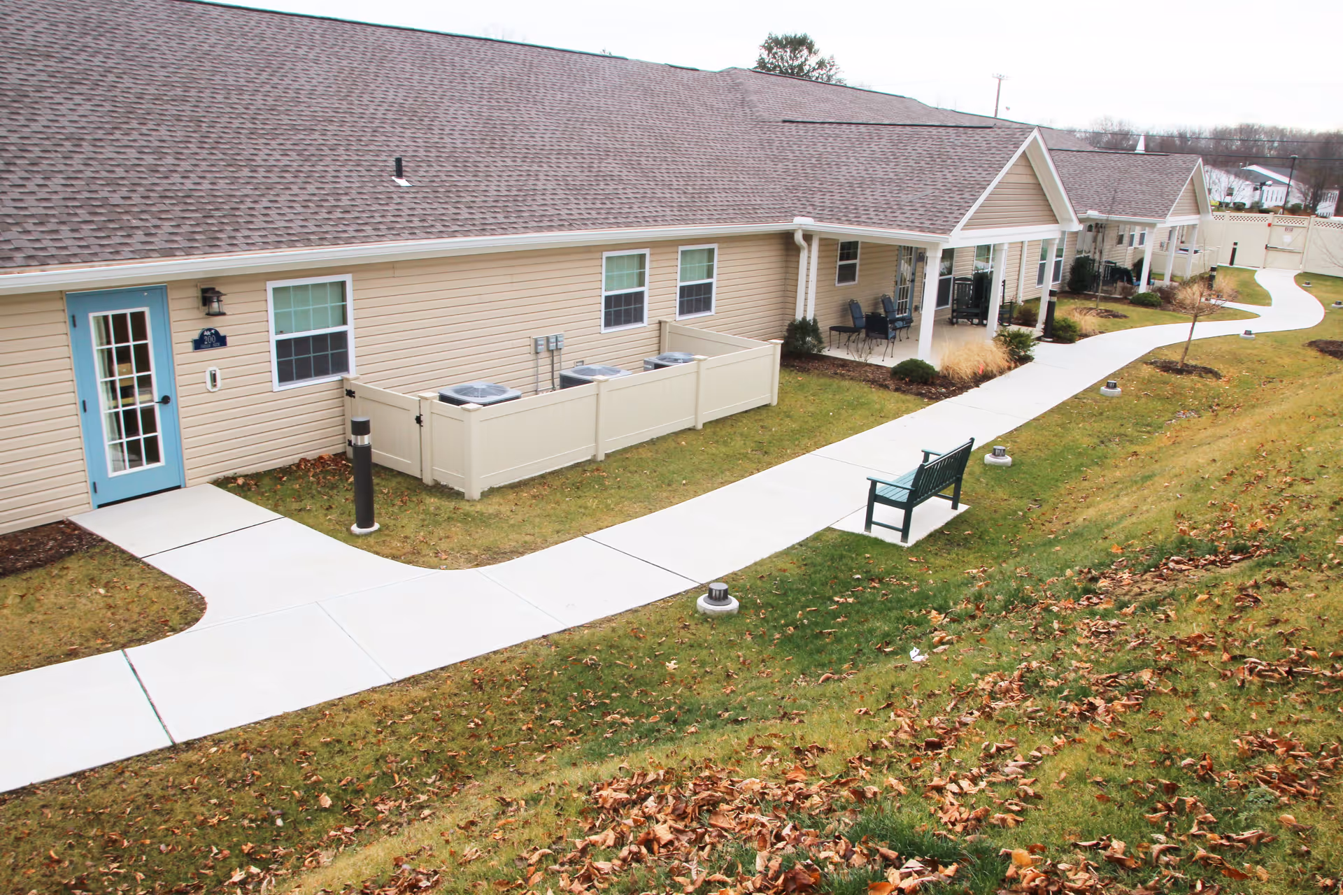 Exterior view of a single-story senior living facility building with beige siding and a brown shingled roof. A paved walkway curves along the front of the building, passing by a green bench and small outdoor seating areas. The lawn is partially covered with fallen leaves, and there are small outdoor lights along the walkway.