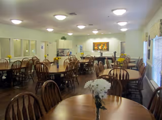 A bright dining room with multiple round wooden tables and chairs, small floral centerpieces, and a serving area/kitchenette at the back.