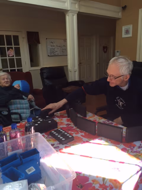 Two elderly individuals sitting at a table playing a game with large domino pieces in a cozy room with beige walls and white pillars. One person is reaching out to place a domino piece while the other watches. The table is covered with a colorful floral tablecloth and various items including water bottles and containers are visible.