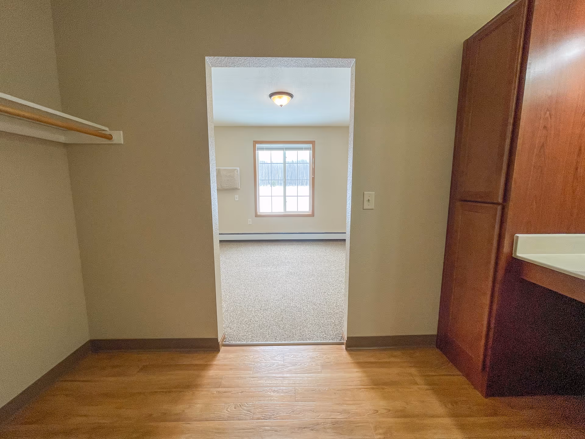 View from a small room with wooden flooring and a closet rod on the left, looking through a doorway into a carpeted room with a window letting in natural light. There is a wooden cabinet and countertop on the right side of the small room.