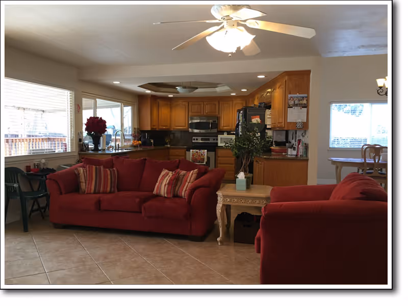 Interior view of a living area with two red sofas and a wooden side table with a plant and tissue box. In the background, there is a kitchen with wooden cabinets, a stove, microwave, and refrigerator. Large windows allow natural light to fill the space, and a ceiling fan with lights is mounted above.