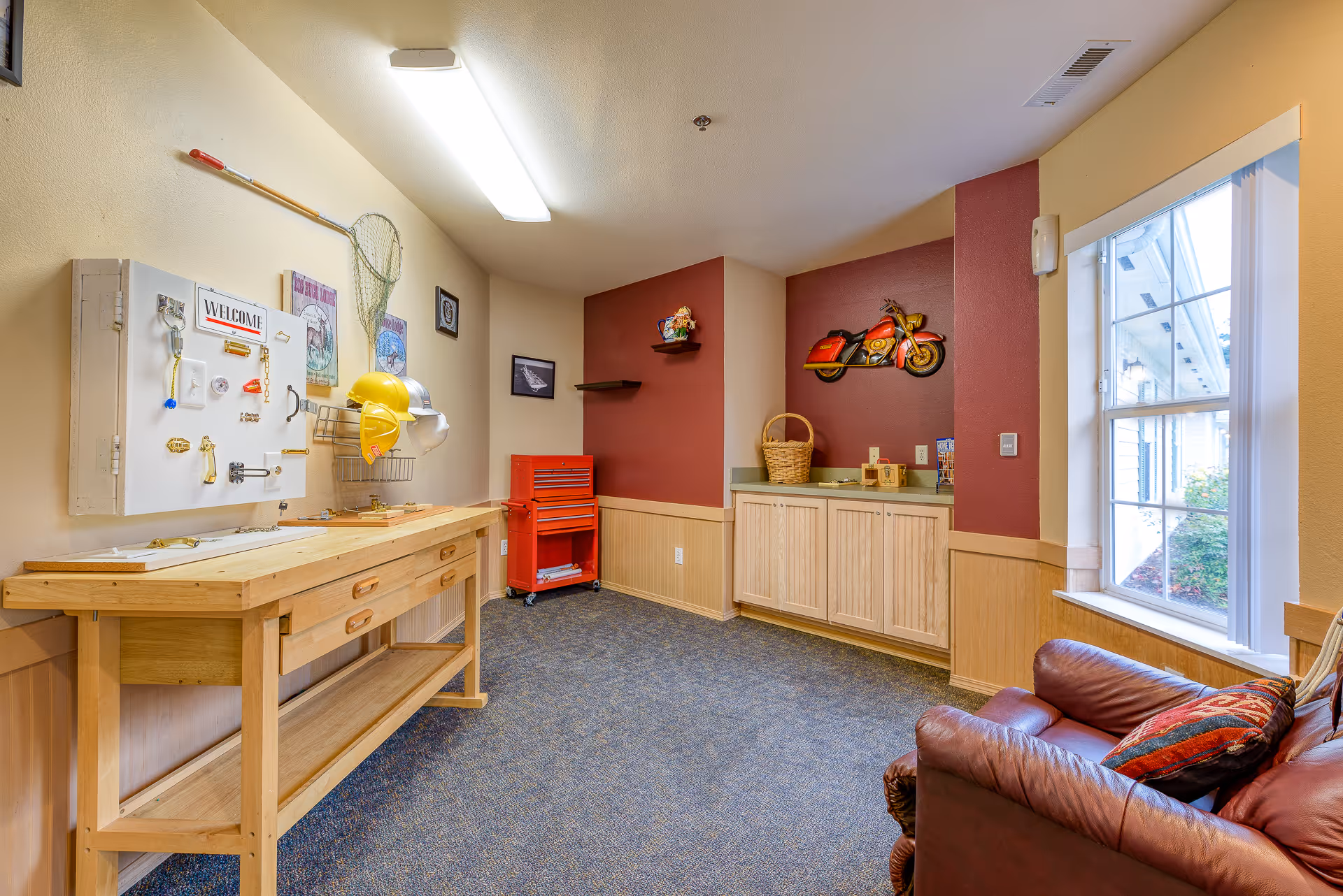 A well-lit interior room with a wooden workbench on the left side featuring various locks and hardware for tactile engagement. Above the workbench are two yellow hard hats and a fishing net hanging on the wall. The back wall is painted red and decorated with a red motorcycle wall art and a small shelf holding a decorative item. Below the shelf are wooden cabinets with a basket on top. On the right side, there is a large window letting in natural light and a brown leather armchair with a colorful pillow. The floor is carpeted in a blue pattern.