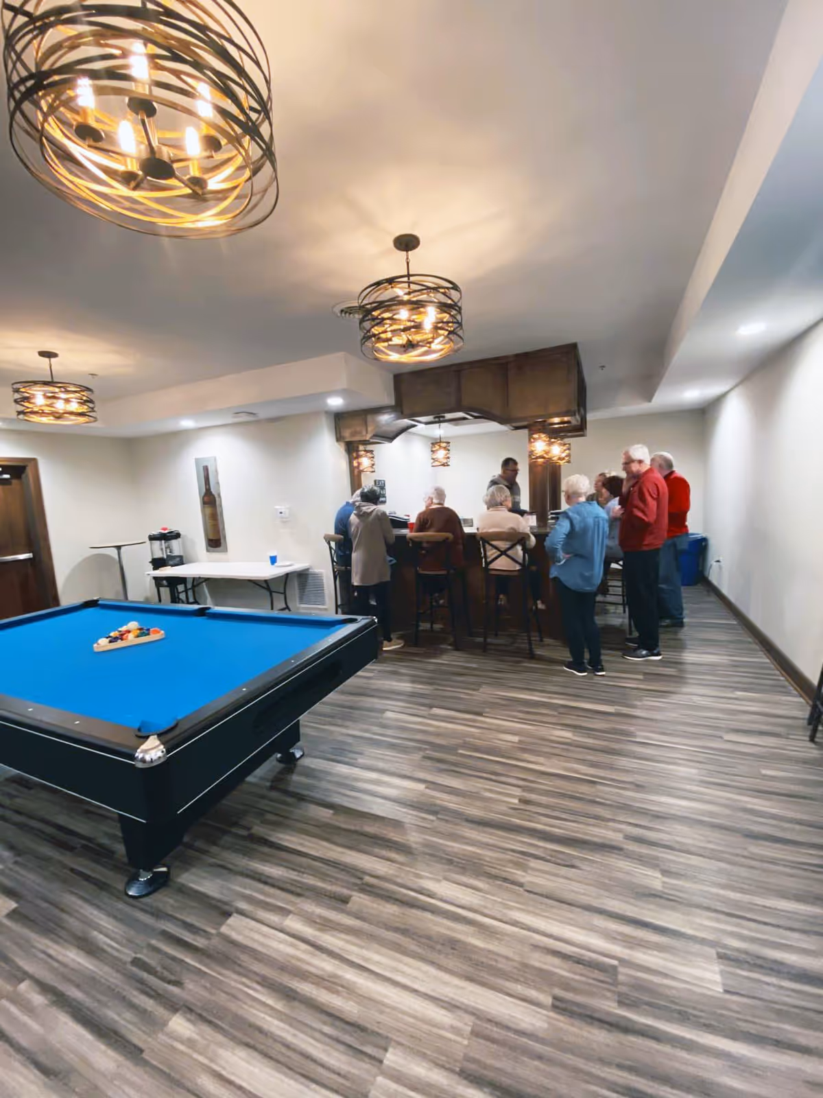 A group of elderly people gathered around a bar area in a recreational room with a pool table in the foreground. The room has modern light fixtures, wood flooring, and neutral-colored walls.