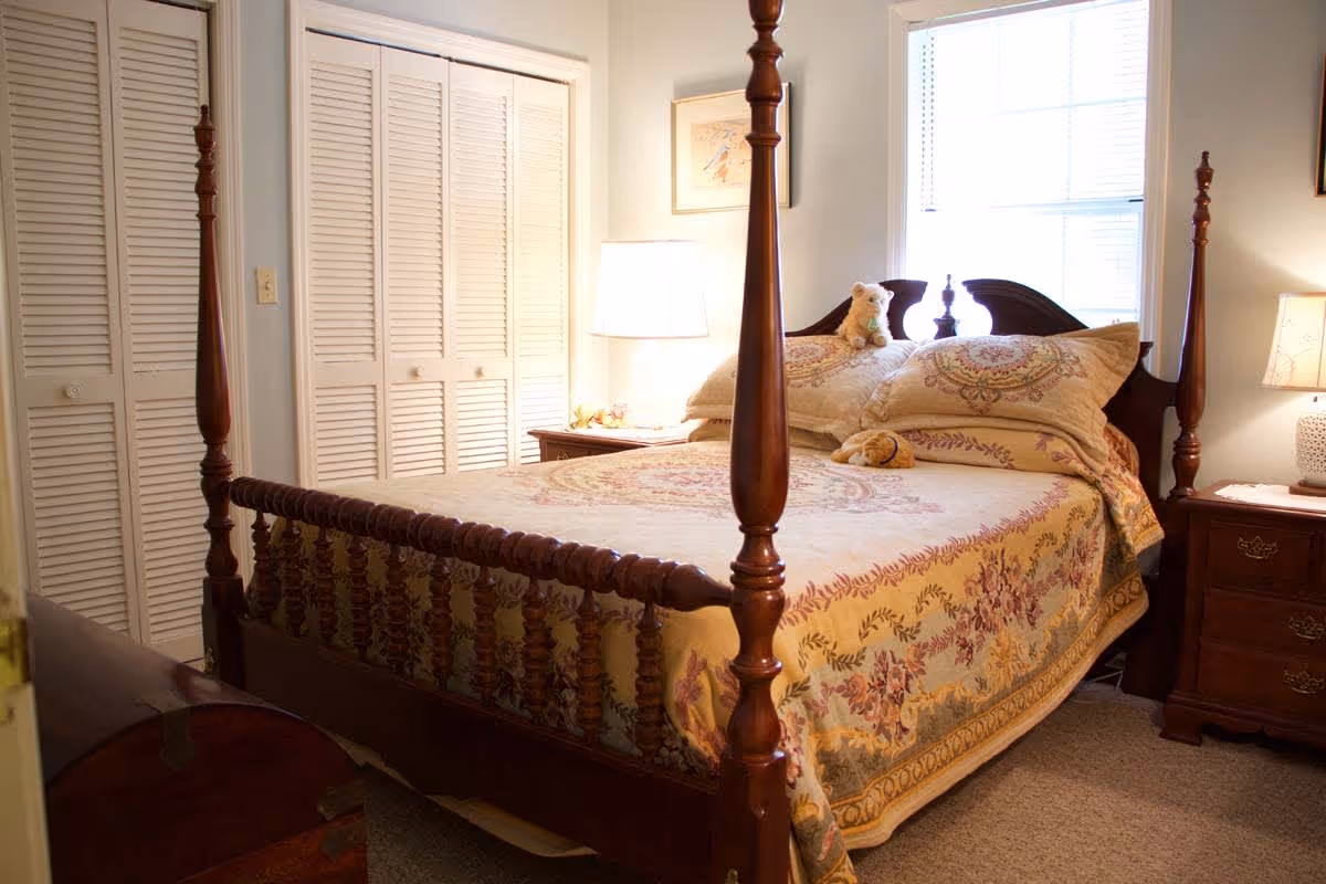 A cozy bedroom with a wooden four-poster bed adorned with a floral patterned bedspread and matching pillows. There are two stuffed animals placed on the bed. The room has a window with blinds letting in natural light, two wooden nightstands with lamps, and a closet with louvered doors.