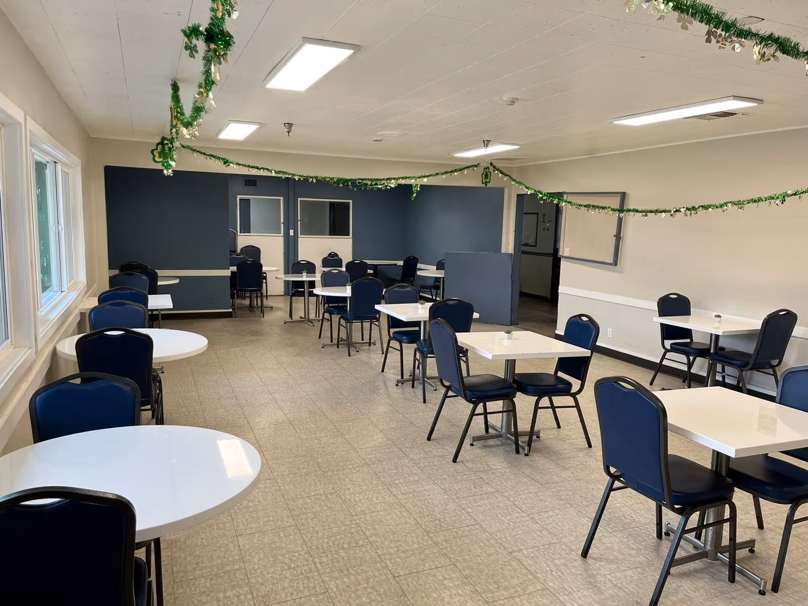 A dining room with multiple white tables and blue chairs arranged neatly. The room has a tiled floor, white walls with a dark blue accent wall at the far end, and fluorescent ceiling lights. Green garlands with shamrock decorations hang across the ceiling. Large windows line one side of the room.