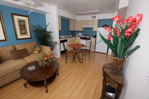 A cozy living area with a beige sofa adorned with patterned cushions, a round wooden coffee table with a floral arrangement, and a small dining table with two chairs. The background features a kitchen with light wood cabinets, a white refrigerator, stove, and microwave. The walls are painted blue and white, and there are framed pictures and a large potted plant adding decoration.