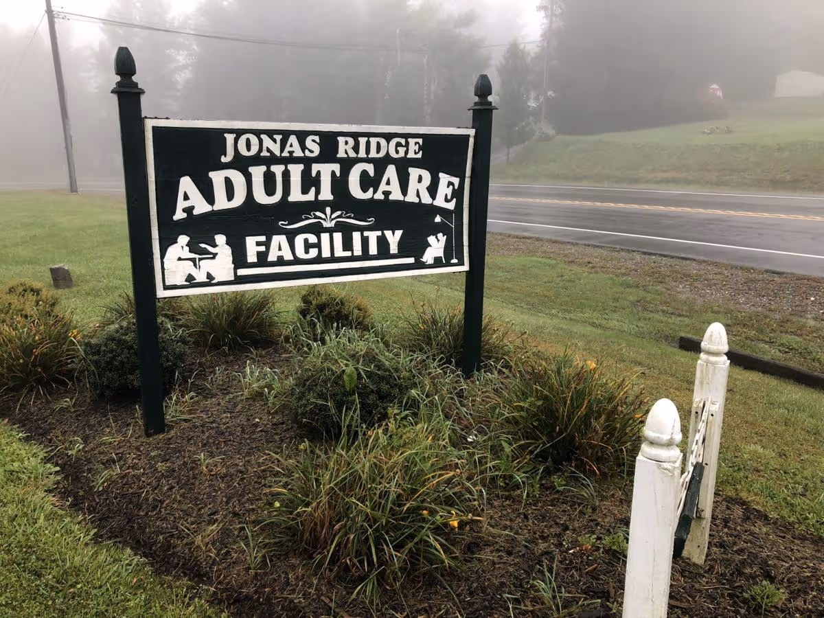A wooden sign for Jonas Ridge Adult Care Facility stands in a landscaped area with grass and plants. The sign is black with white text and illustrations of people sitting and interacting. In the background, there is a road and a foggy, tree-lined area.
