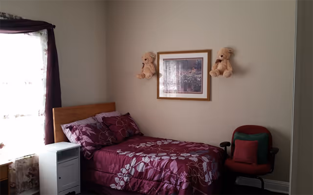 A small bedroom with a single bed covered in a purple floral bedspread and matching pillows. Two teddy bears are mounted on the wall above the bed, flanking a framed picture. There is a white nightstand next to the bed and a red cushioned chair with green and red pillows beside it. A window with sheer floral curtains and dark purple drapes is on the left side of the room.