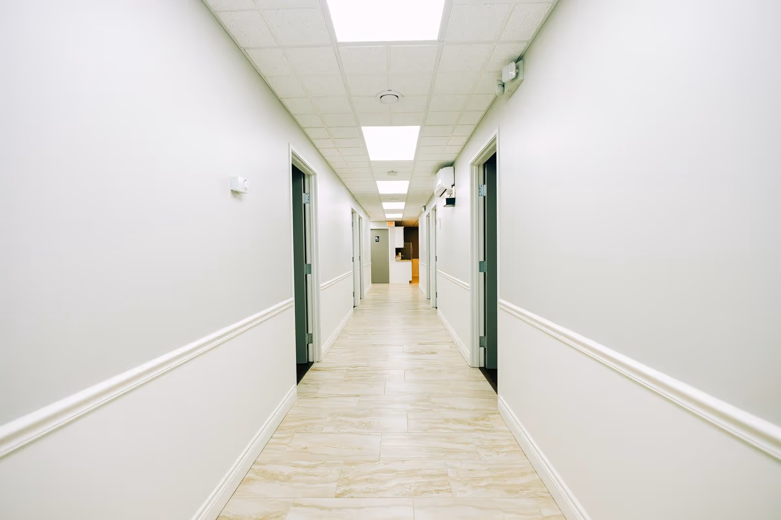 A long, well-lit hallway with white walls and light-colored tiled flooring. Several open doorways line both sides of the corridor, and the ceiling has recessed lighting panels.