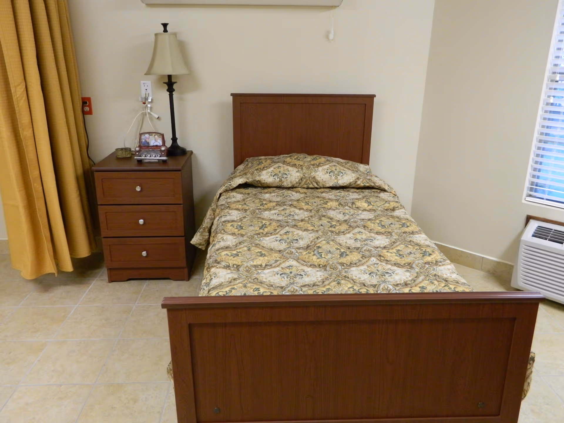 A single bed with a patterned bedspread in a room. Next to the bed is a wooden nightstand with three drawers, a table lamp, a framed photo, and some small items. There is a window with blinds and an air conditioning unit below it. Yellow curtains hang on the left side of the image.