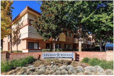 Exterior view of Studio Royale assisted living facility showing a two-story building with beige and brown walls, surrounded by trees and landscaping. A brick wall in front displays a sign with the facility's name, Studio Royale.