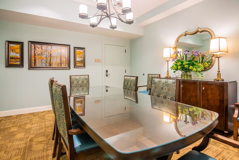 A well-lit dining room with a large polished wooden table surrounded by upholstered chairs with patterned backs. On one wall, there are three framed paintings of autumn trees. Opposite the paintings, there is a wooden sideboard with two table lamps and a large decorative mirror above it. A vase with a colorful floral arrangement sits on the sideboard. The room has light green walls and a carpeted floor.