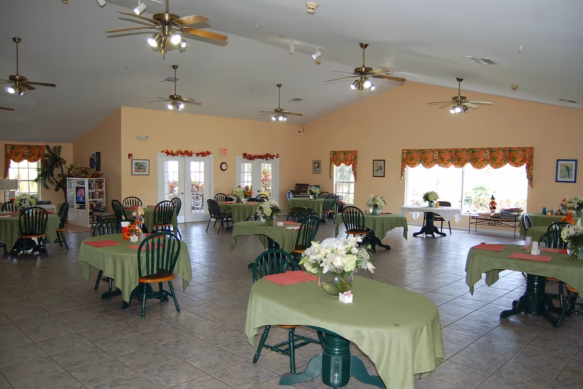 A spacious dining room with multiple round tables covered in green tablecloths, each adorned with a vase of white flowers. The room has tiled floors, ceiling fans with lights, large windows with floral valances, and double glass doors. There are green wooden chairs around the tables and a television and bookshelf in the background.
