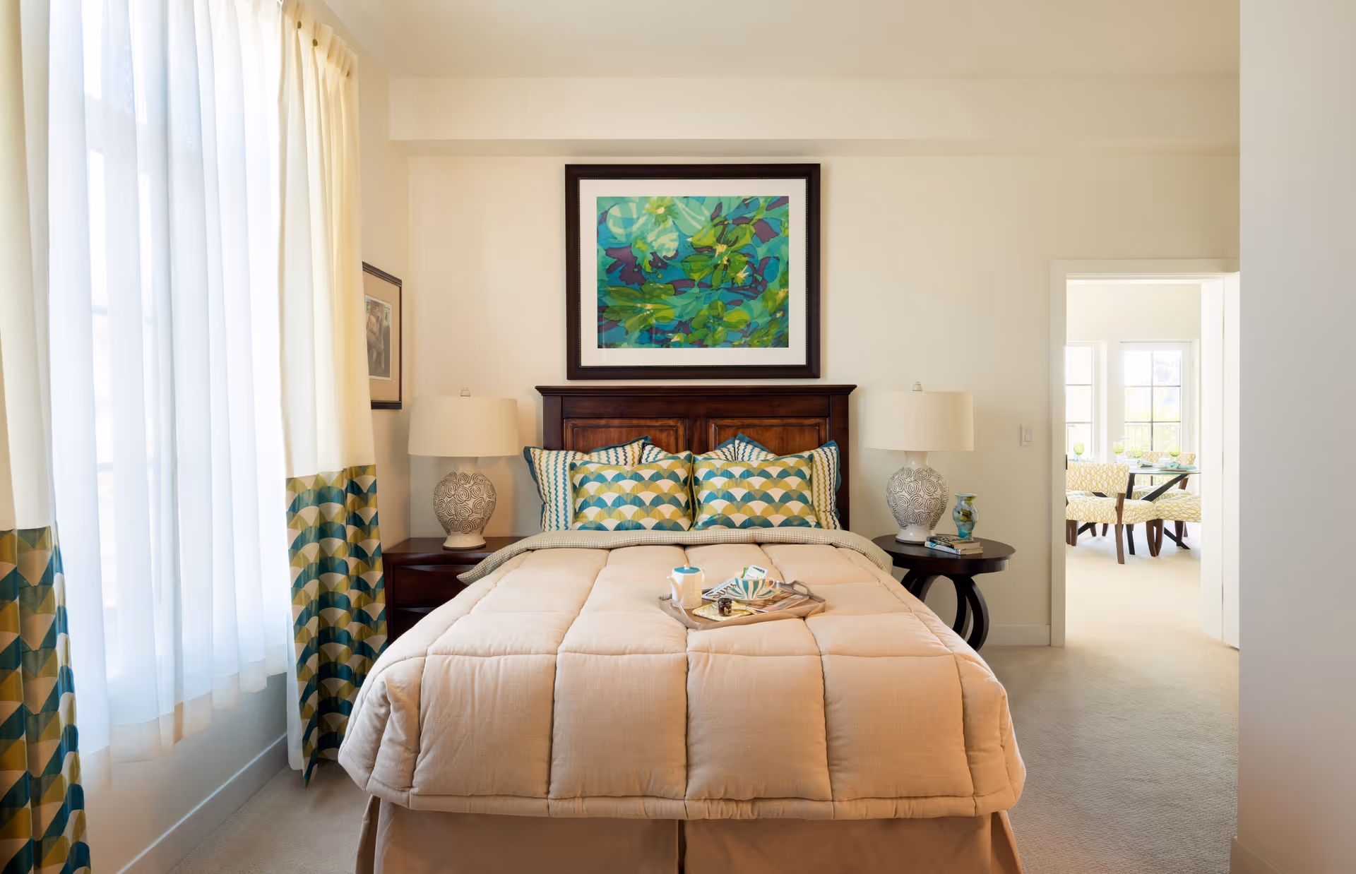 Well-lit bedroom with a neatly made bed, patterned pillows, bedside tables with lamps, and an open doorway to a bright sitting area.