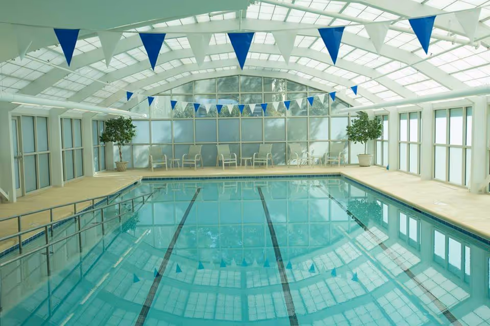 Indoor swimming pool with clear blue water, surrounded by a beige deck. The pool area is enclosed by large windows and a white arched ceiling with blue triangular flags hanging across. There are several chairs and potted plants along the far wall.