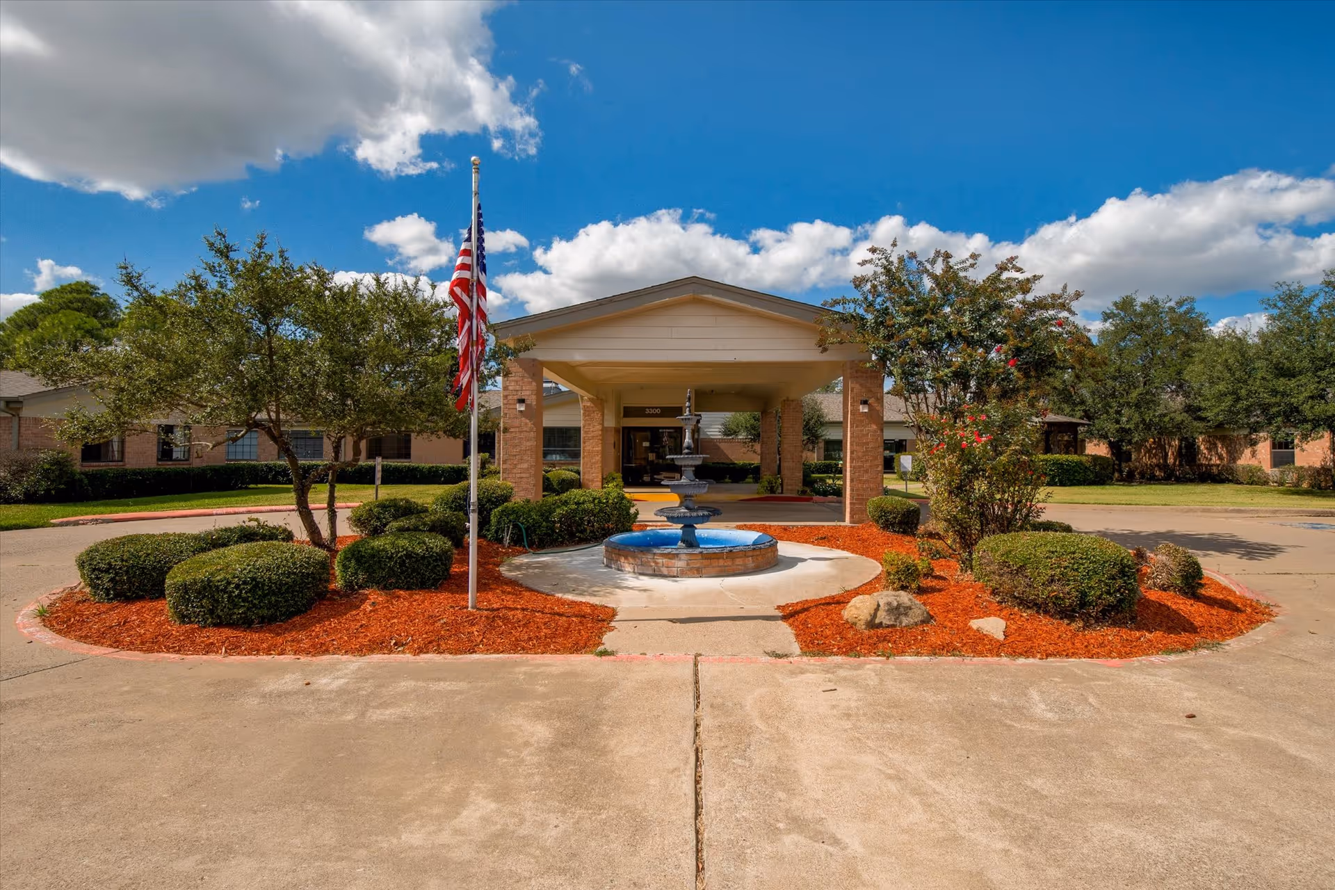 Front exterior view of Legacy at Corsican facility showing a covered entrance with a three-tiered fountain in the center, surrounded by neatly trimmed bushes and trees, an American flag on a flagpole, and a bright blue sky with scattered clouds.