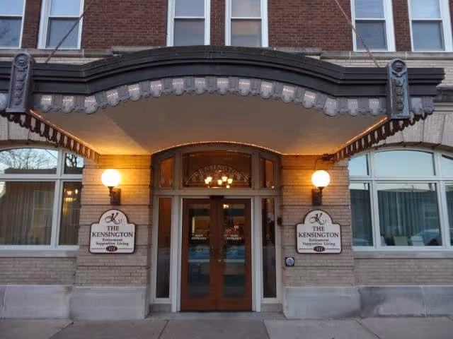 Entrance to The Kensington retirement and supportive living facility with double wooden doors under a decorative awning, flanked by two illuminated wall lamps and signs displaying the facility's name.