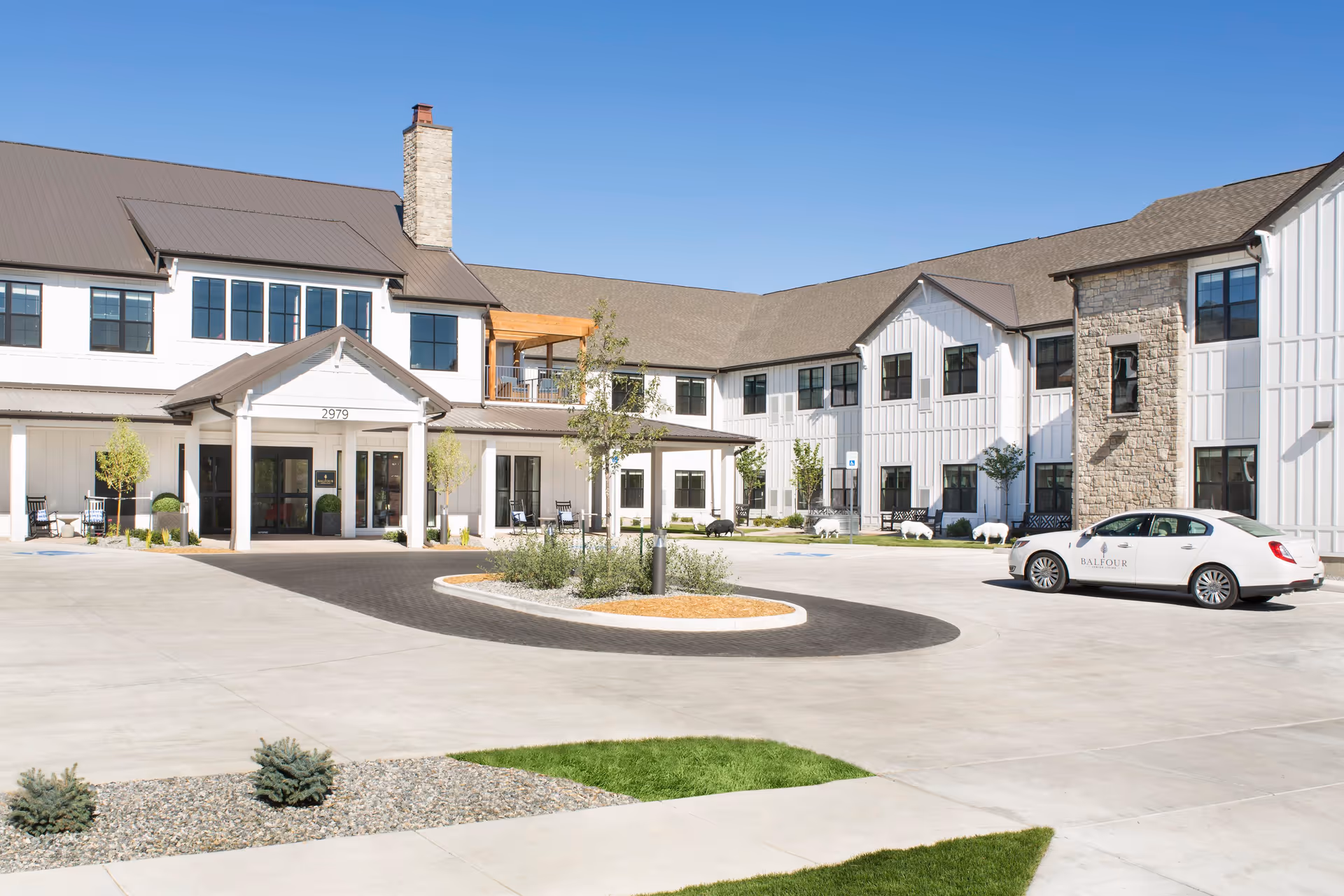 Exterior view of Balfour Central Park senior living facility showing a large, modern two-story building with white siding, stone accents, and a brown roof. There is a circular driveway with a landscaped island in the center, a white car parked on the right side with the Balfour logo, and clear blue sky overhead.