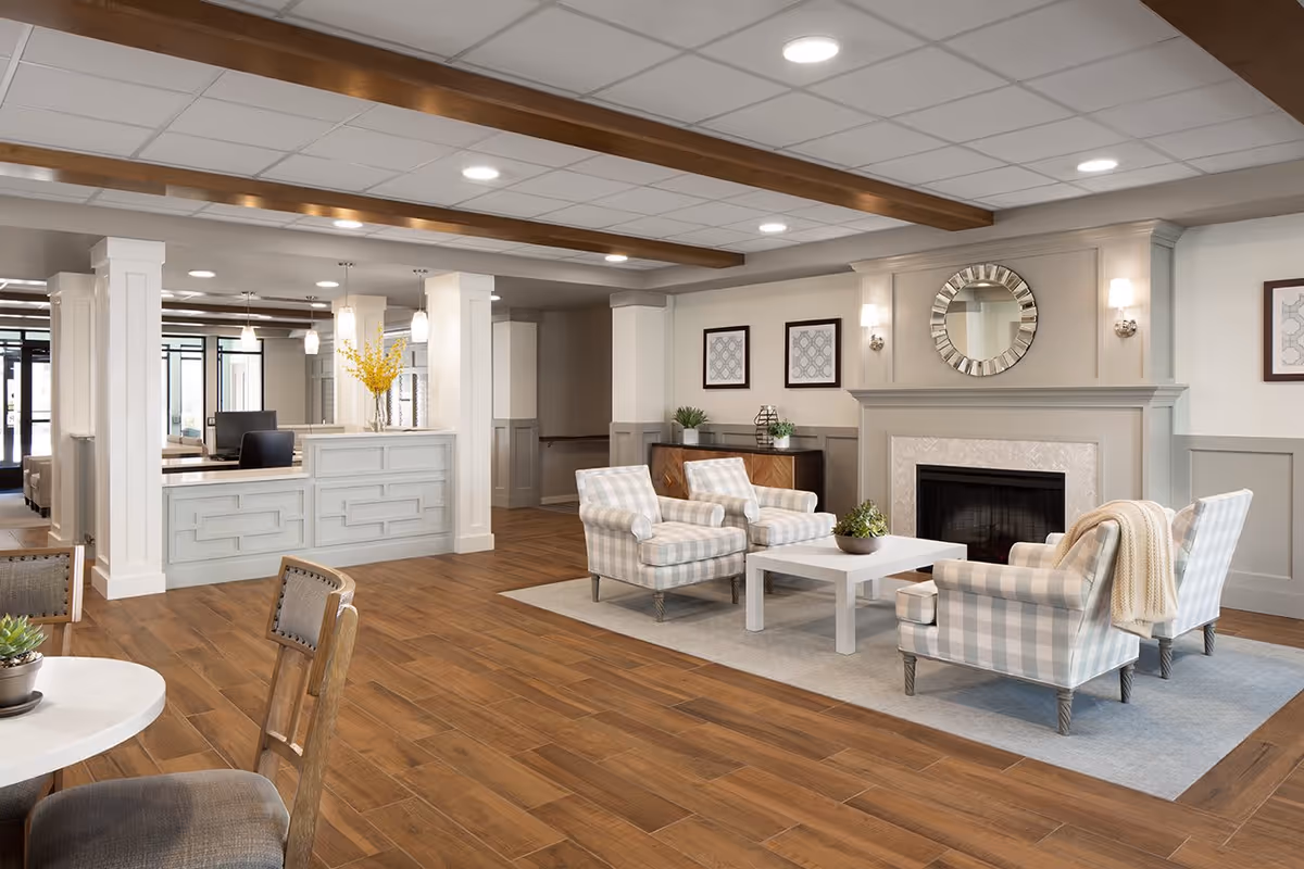 A spacious and well-lit senior living facility common area featuring a seating arrangement with four plaid armchairs around a white coffee table on a light gray rug. Behind the seating area is a fireplace with a decorative round mirror above it and two framed artworks on the wall. To the left is a reception desk with pendant lights and a vase with yellow flowers. The room has wooden flooring and ceiling beams, with neutral-colored walls and modern lighting.