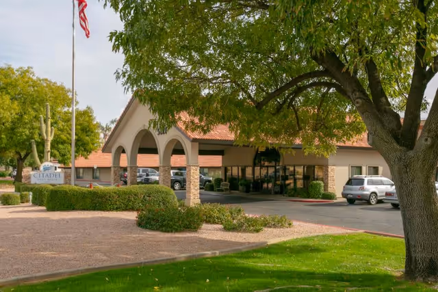 Exterior view of Citadel Post Acute facility showing the entrance with arched columns, a flagpole with an American flag, parked cars, and surrounding trees and landscaping.