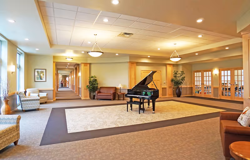 A spacious and well-lit common area in a senior living facility featuring a grand piano on a large patterned rug in the center. The room has comfortable seating with armchairs and sofas, potted plants, and large windows allowing natural light. There are wooden framed glass doors on one side and a hallway leading to other rooms.