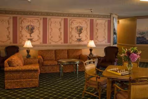 Cozy senior living lounge with patterned wallpaper, sofas, lamps, and a round table.