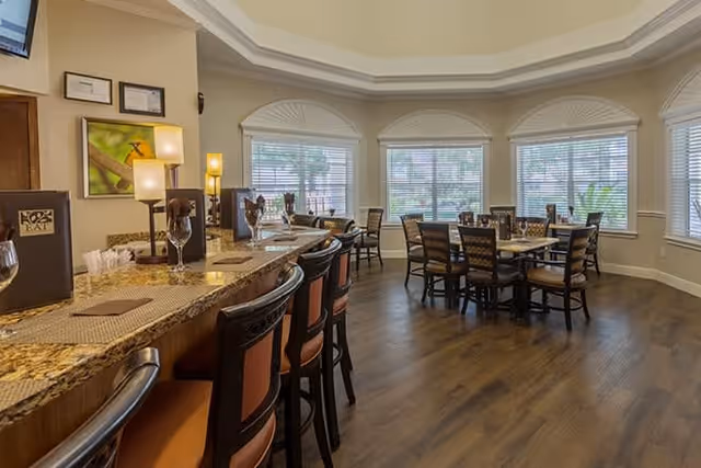 Interior view of a dining area in a senior living facility with a granite countertop bar lined with chairs on the left and several dining tables with chairs near large windows on the right. The room has wooden flooring, soft lighting, and neutral-colored walls with decorative molding on the ceiling.