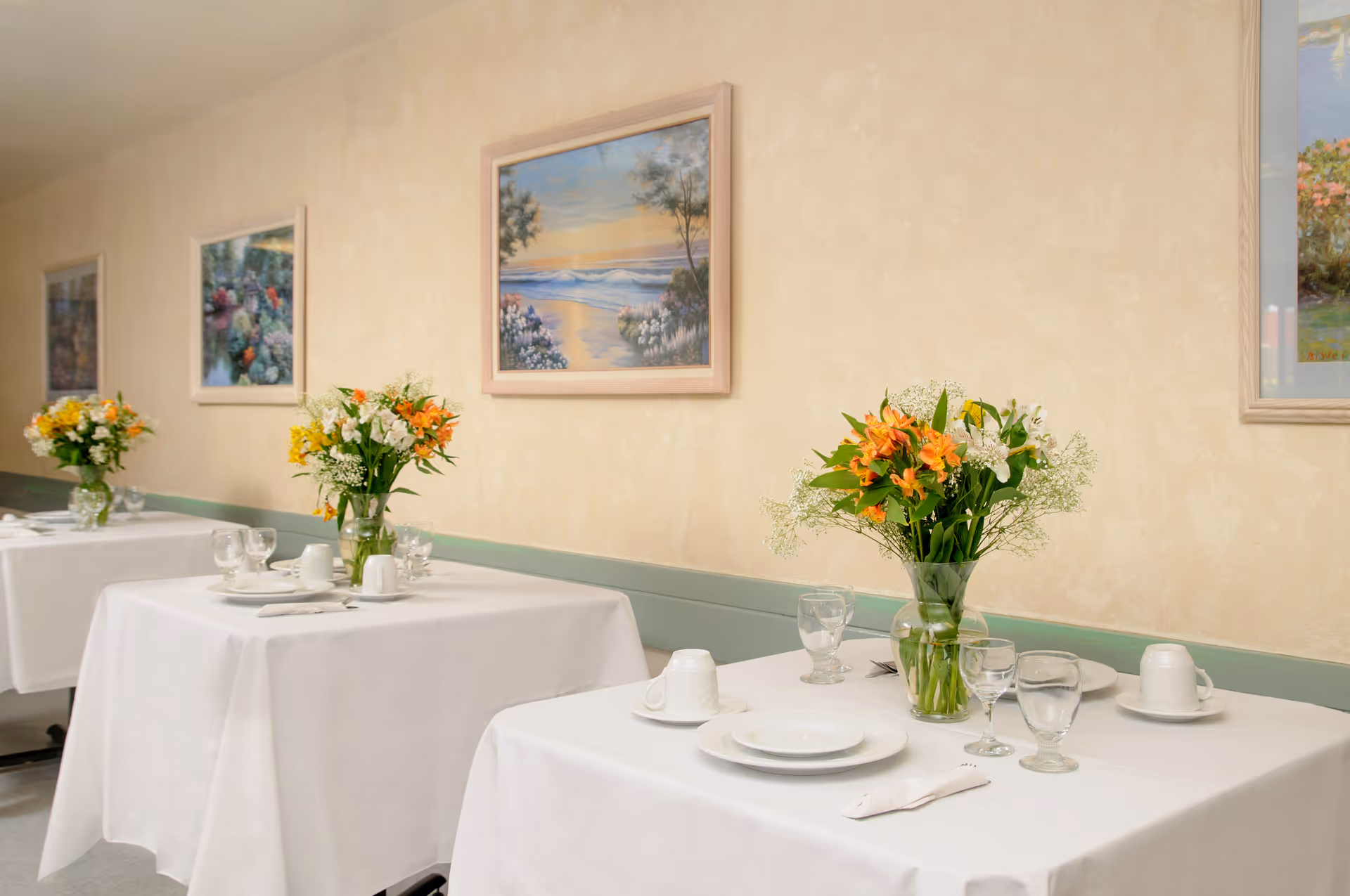 Tables in a dining room set with white tablecloths, place settings, and vases of flowers against a wall with framed paintings.