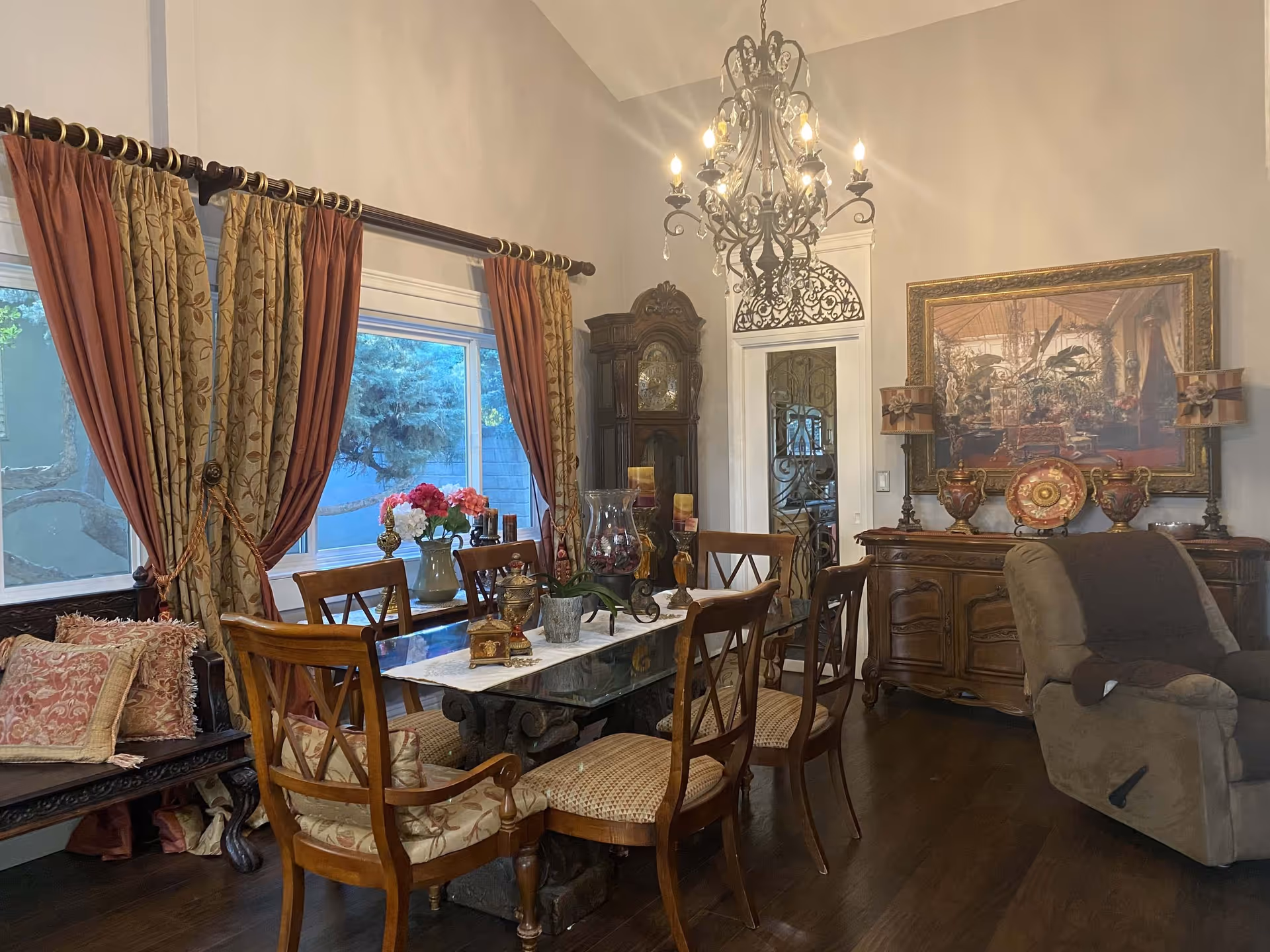 Ornately furnished dining room with a wooden table and chairs, chandelier, large window with drapes, and a sideboard.