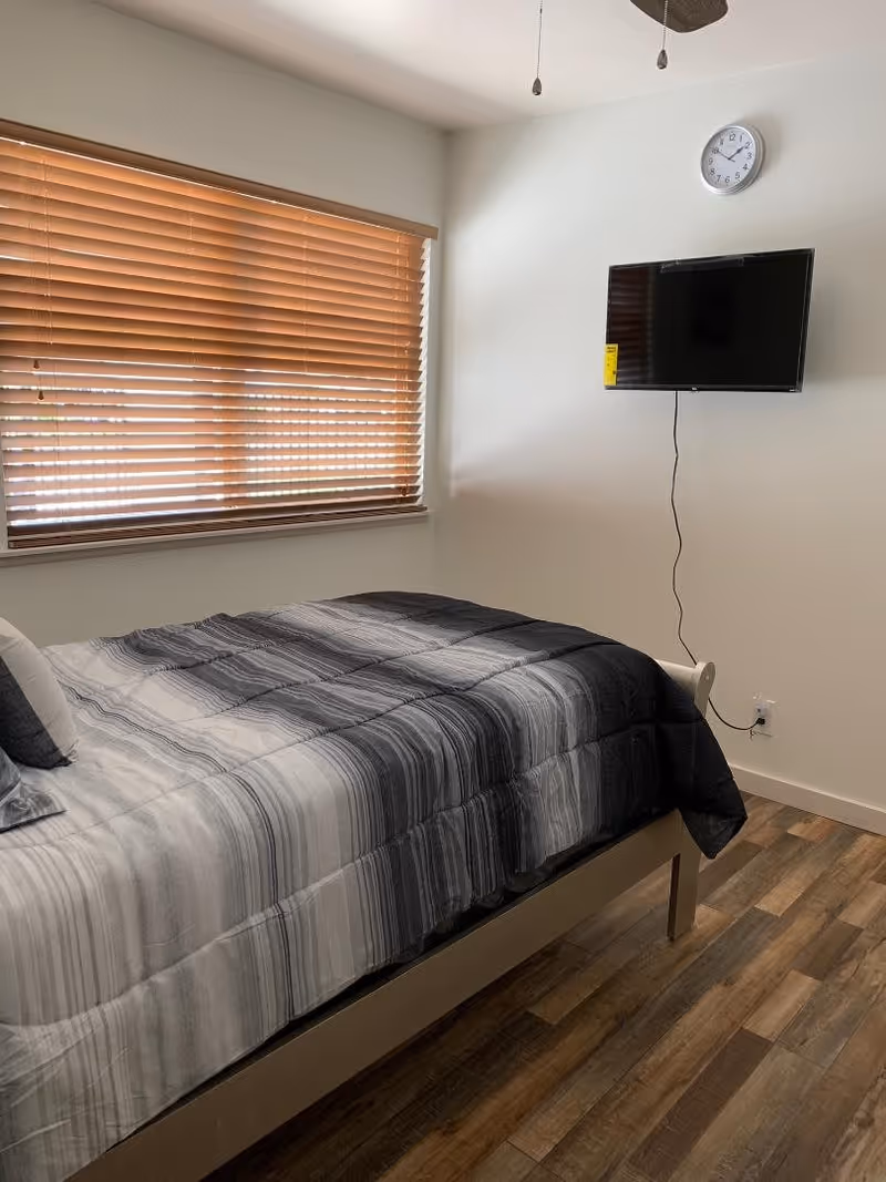 A bedroom with a bed covered in a gray and black striped comforter, a pillow, a window with wooden blinds, a wall-mounted flat-screen TV, and a clock on the wall above the TV.