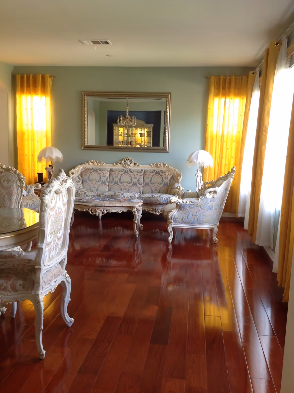 A living room with polished wooden floors, ornate vintage-style upholstered furniture including a sofa, armchair, and dining chairs. The walls are painted light green, with two yellow curtains on windows letting in natural light. A large mirror hangs on the wall above the sofa, reflecting a chandelier and a cabinet.