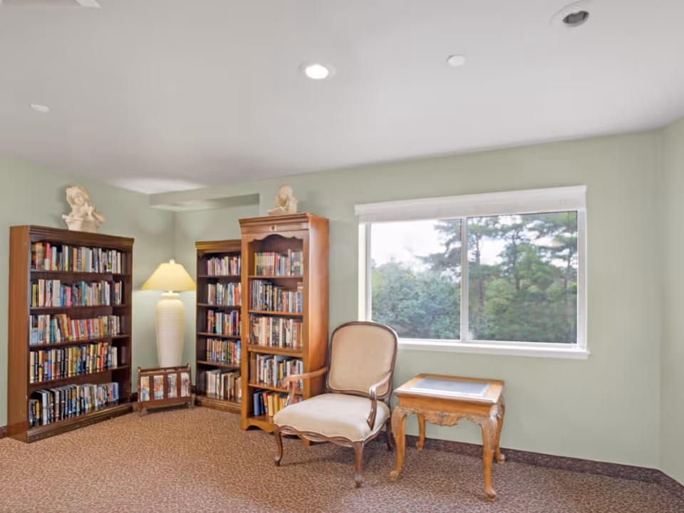 A cozy reading nook in a senior living facility featuring two wooden bookshelves filled with books, a tall white lamp between them, a cushioned armchair, and a small wooden side table. A large window shows green trees outside, and the walls are painted light green.