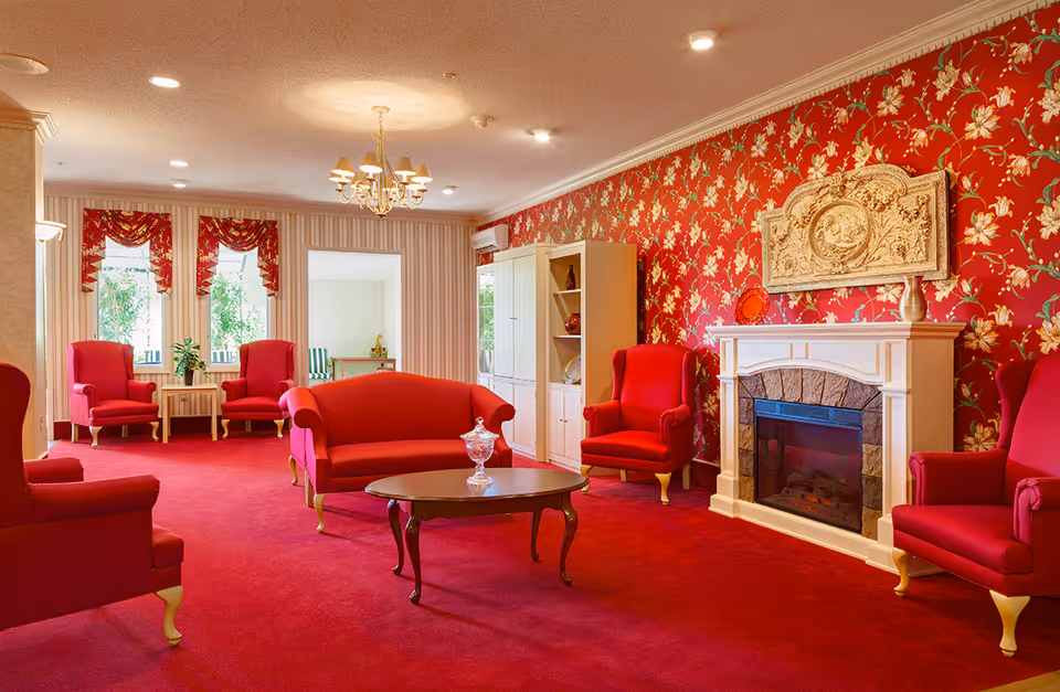 Bright traditional lounge with red upholstered chairs and sofas, patterned red wallpaper, and a white fireplace.