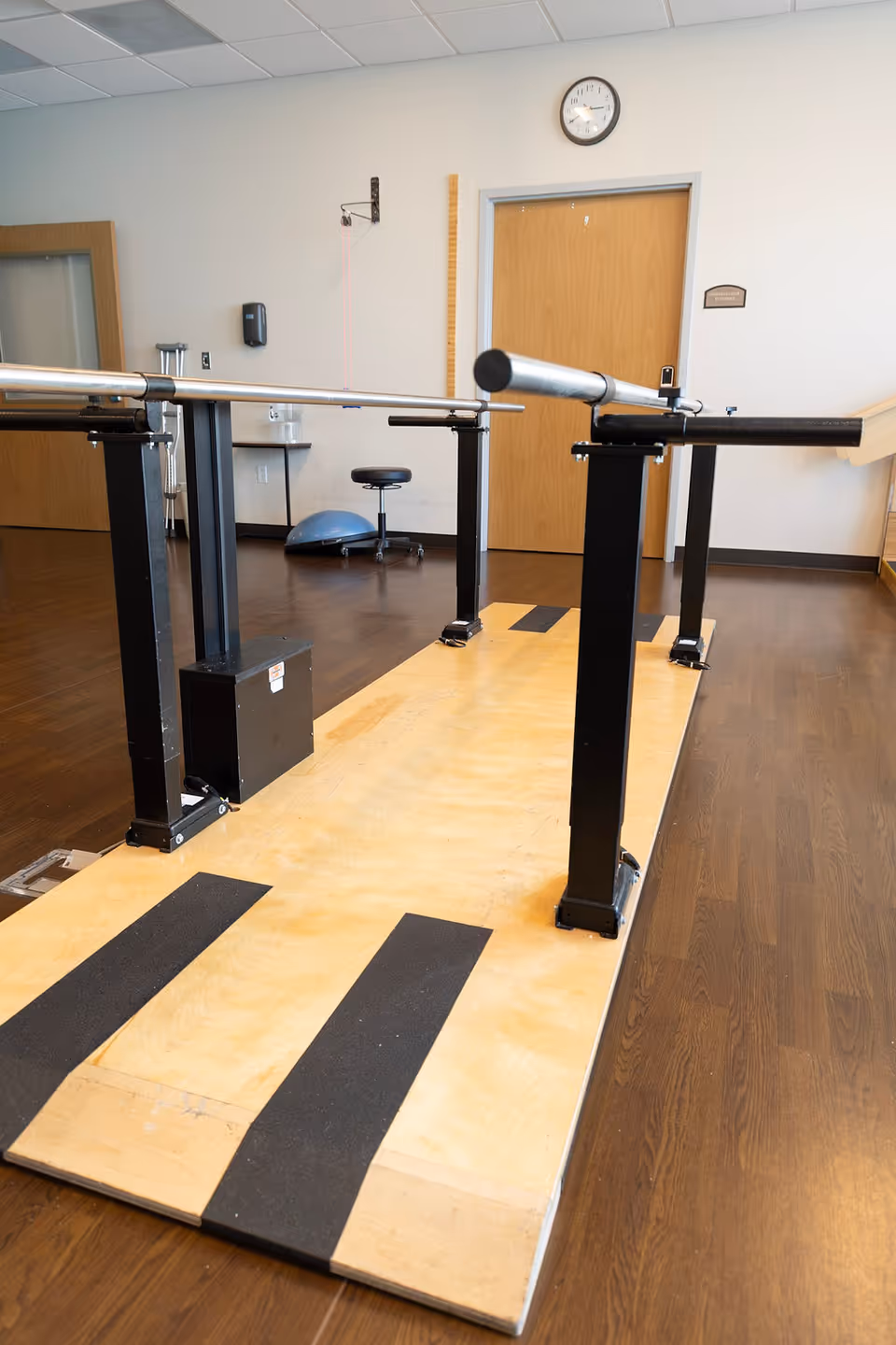Physical therapy room with parallel bars mounted on a wooden platform for walking exercises, a stool, a balance dome, and a clock on the wall above a closed wooden door.