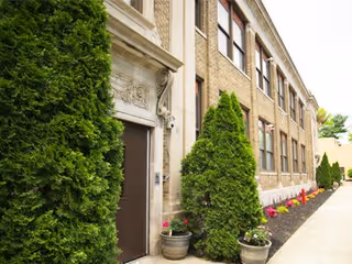 Exterior view of a brick building with multiple windows, a brown door framed by decorative stonework, and a sidewalk lined with potted plants and neatly trimmed evergreen bushes.
