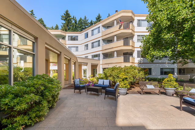 Outdoor patio area at Cristwood Park featuring seating arrangements with chairs and a small table, surrounded by green bushes and trees, with a multi-story building in the background under a clear blue sky.