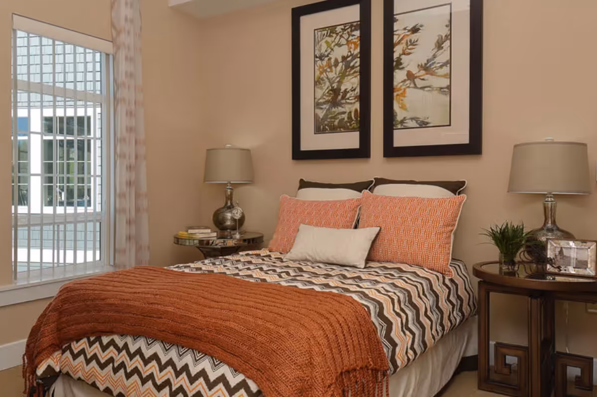 Cozy bedroom with a patterned bedspread and orange throw, two bedside tables with lamps, framed artwork above the bed, and a window with shutters.