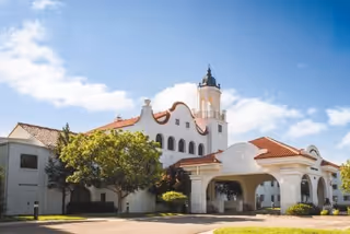 Exterior view of a large, white building with Spanish-style architecture featuring a red tile roof, arched windows, and a tower with a dome. The building is surrounded by green trees and bushes under a blue sky with some clouds.
