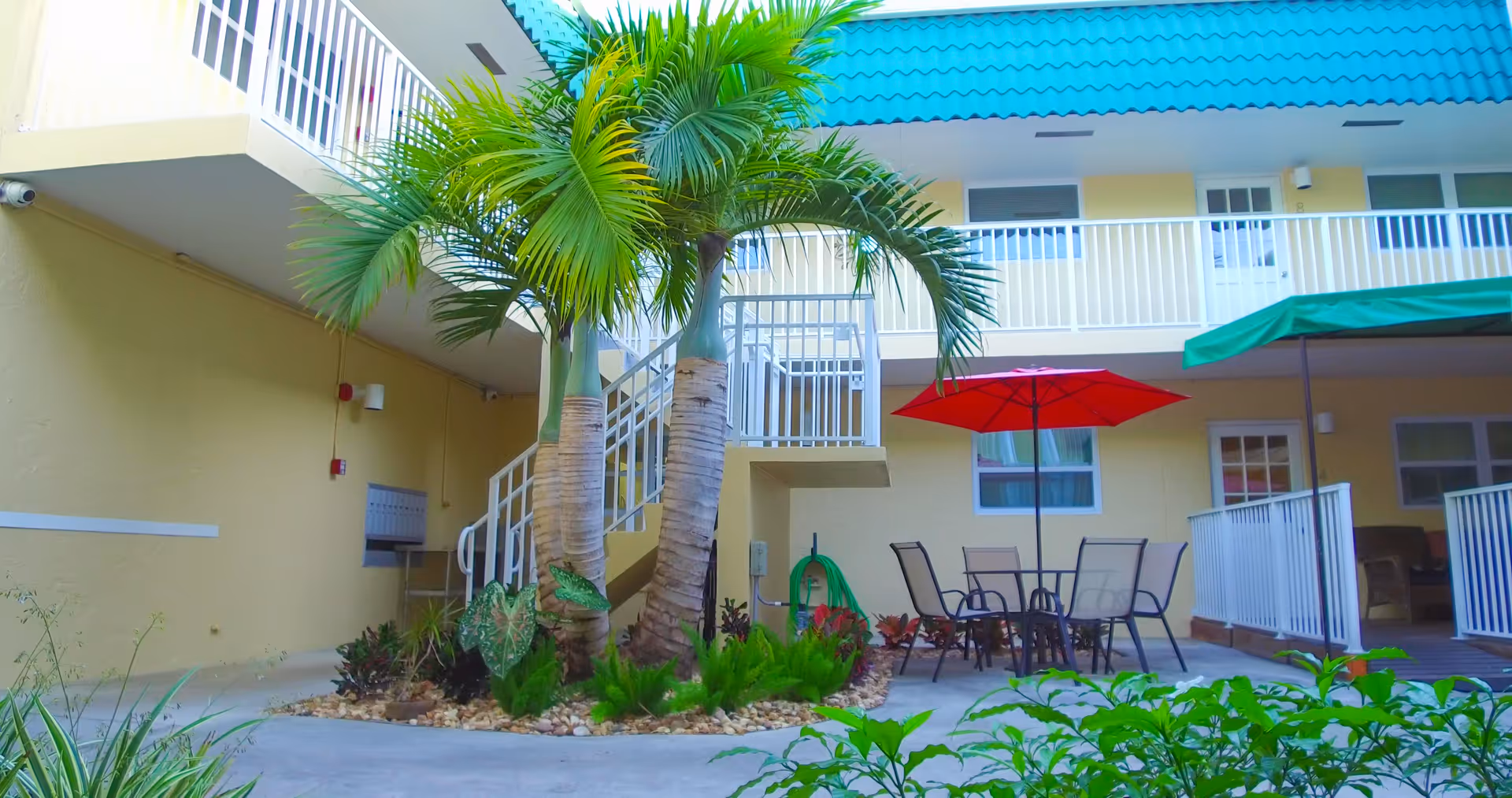 Outdoor courtyard area with two palm trees surrounded by small plants and rocks. There is a patio table with a red umbrella and several chairs. The building surrounding the courtyard has yellow walls, white railings, and a blue roof. A green umbrella is also visible on the right side.