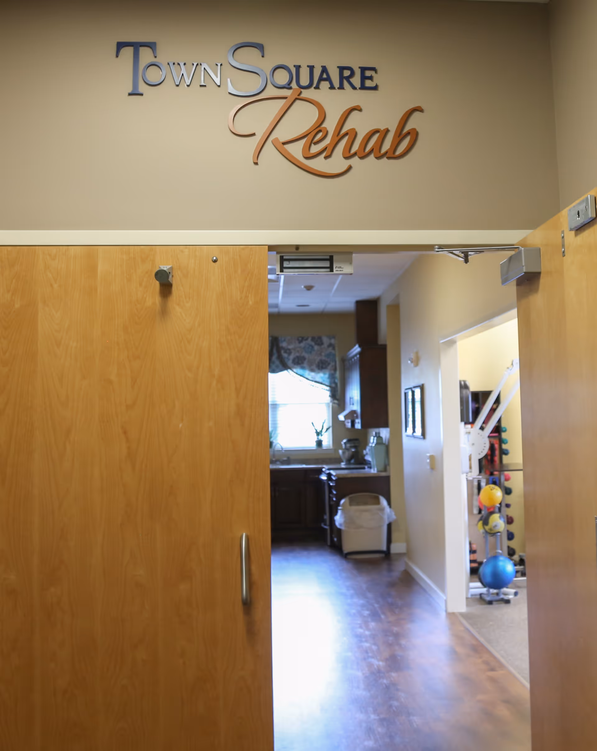 Open double wooden doors leading into a rehabilitation area with a sign above reading 'Town Square Rehab'. Inside, there is a room with exercise equipment including colorful weights and a blue exercise ball, and a kitchen area with a window, cabinets, and a trash bin.