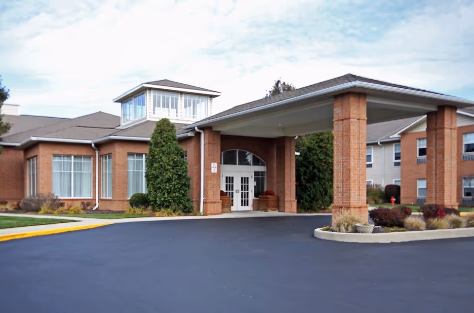 Exterior view of The Waterford at Fairfield senior living facility showing the main entrance with a covered drop-off area supported by brick columns, surrounded by landscaped greenery and a paved driveway.