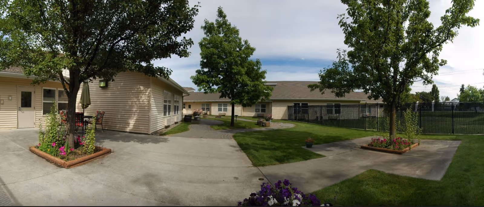 Outdoor courtyard area of a senior living facility with concrete walkways, green grass, flower beds with blooming flowers, several trees, and beige single-story buildings surrounding the space under a partly cloudy sky.