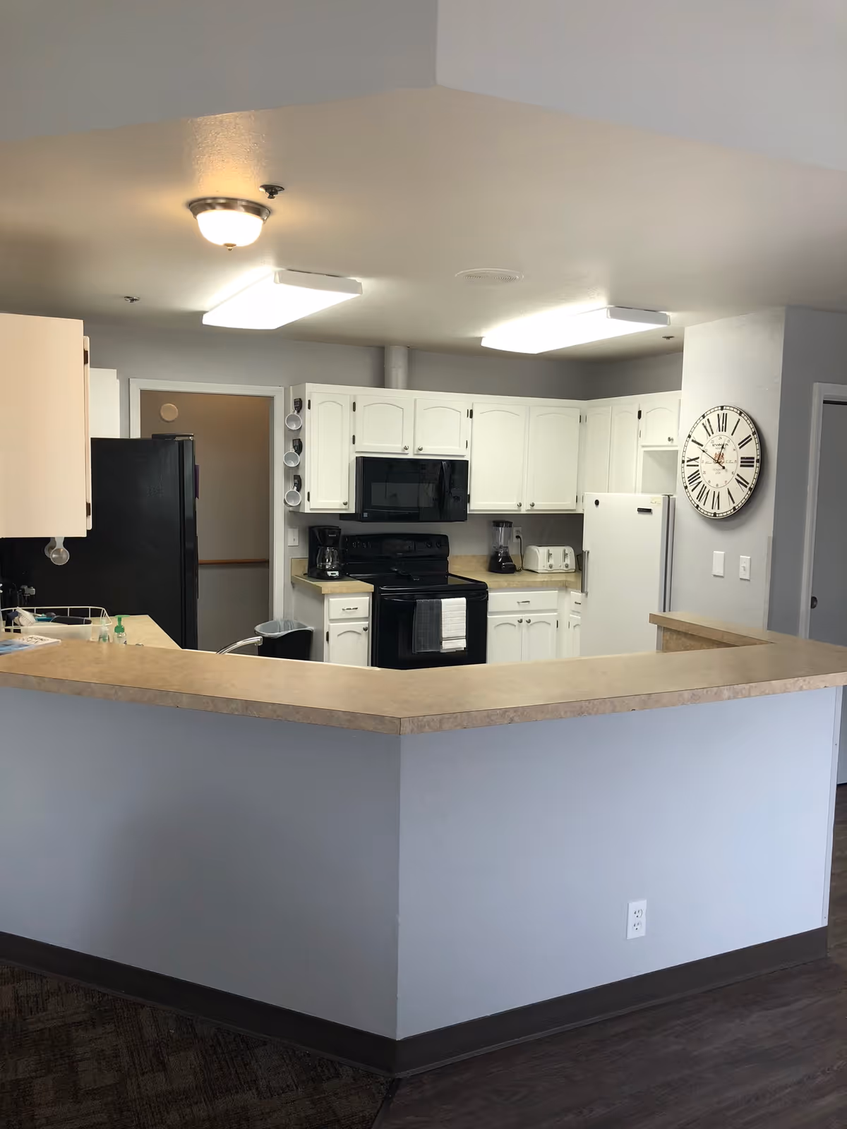 Interior view of a kitchen in an assisted living facility featuring white cabinets, a black stove with a microwave above it, a white refrigerator, a coffee maker, a blender, a toaster, and a large wall clock. The kitchen has a beige countertop with a sink and is well-lit with ceiling lights.