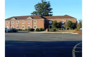 Exterior view of a two-story brick building with multiple windows, a tree in front, and a parking lot with several cars parked. The sky is clear and blue.
