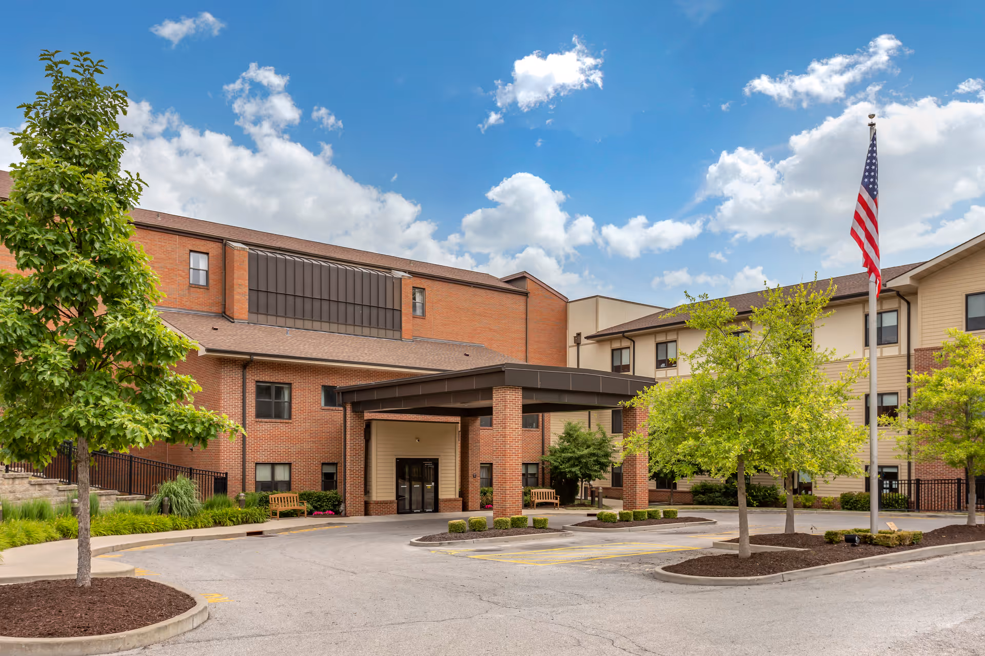 Exterior view of St Paul's Senior Community building with a covered entrance, brick and beige siding, several windows, landscaped trees and bushes, an American flag on a flagpole, and a partly cloudy blue sky.