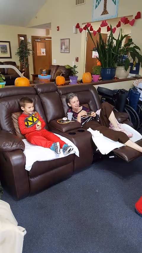 An elderly woman and a young boy sitting on a brown leather reclining sofa in a cozy living room. The woman is reclining with her legs up, holding a plate of food, while the boy is sitting upright eating a snack. The room is decorated with pumpkins, potted plants, and a garland of red hearts hanging on the wall. In the background, there are more sofas, framed pictures, and a wheelchair.