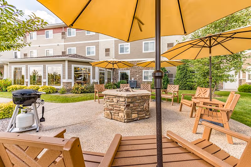 Outdoor patio area at New Perspective Senior Living | Waconia featuring wooden chairs arranged around a stone fire pit, several large yellow umbrellas providing shade, a propane grill, and a multi-story building with windows and landscaping in the background.