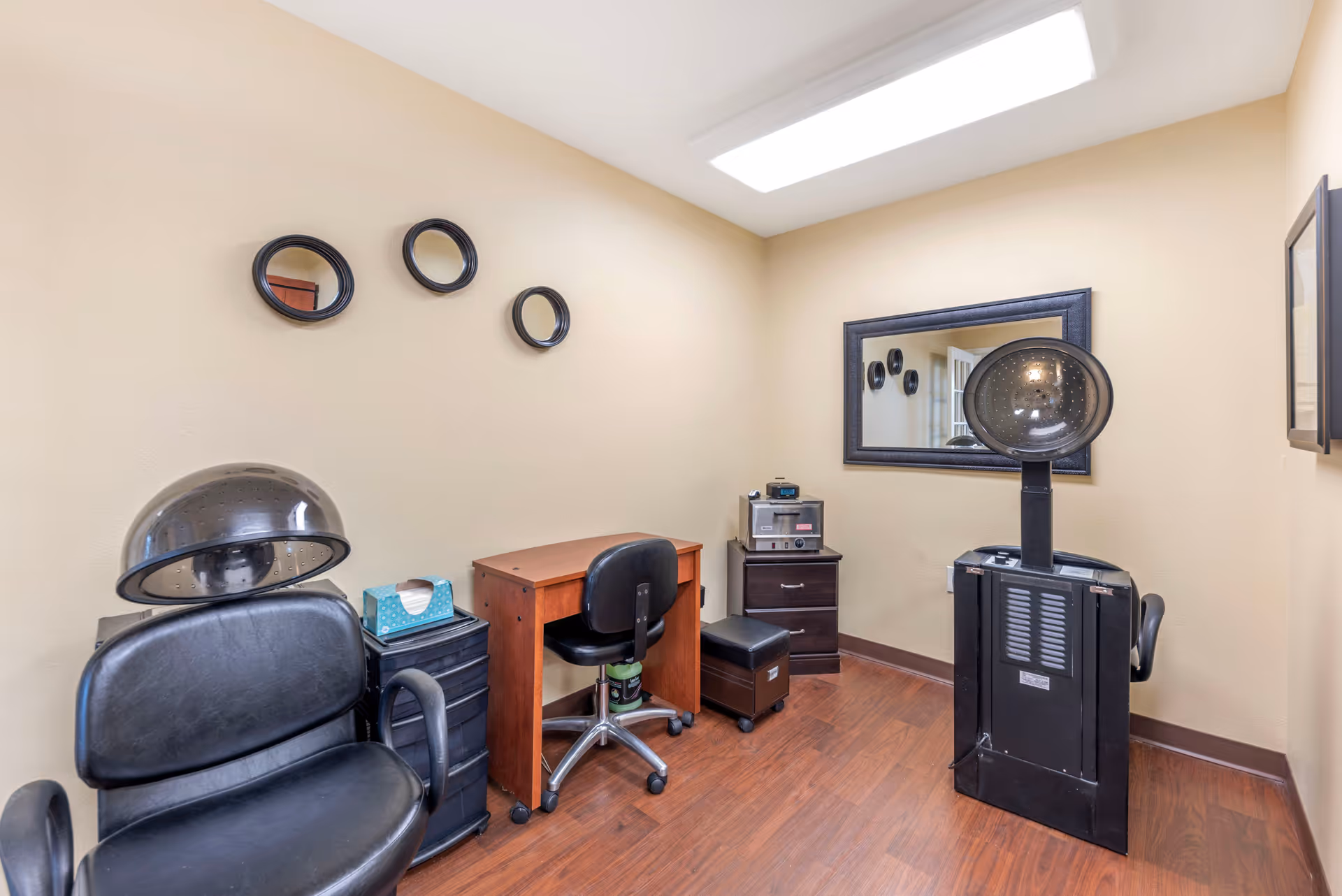 Small salon-style room with two hooded hair dryers, salon chairs, a desk, mirror, and wood flooring.